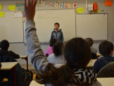 Un collègien se tient debout, au tableau, devant sa classe, une collégienne assise à sa table lève la main