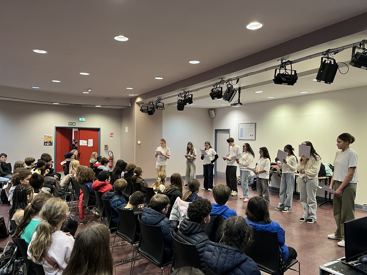 9 élèves se tiennent debout avec des textes à la main devant un public composé d'élèves
