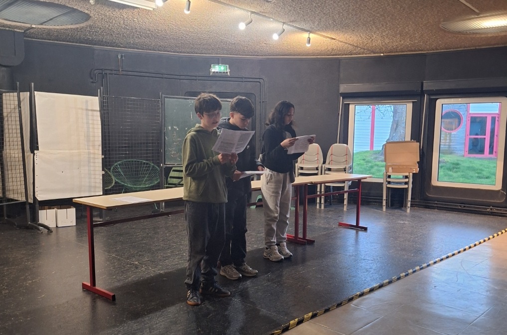 3 élèves se tiennent debout avec un texte à la main dans une grande salle