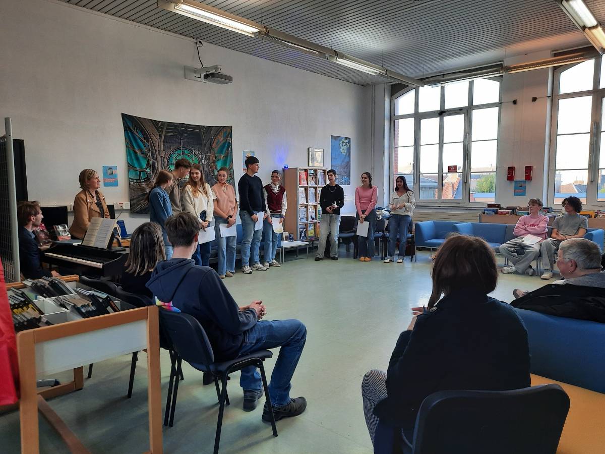 Des élèves lisent des textes dans le CDI devant un public assis autour d'eux, un enseignant les accompagne au clavecin