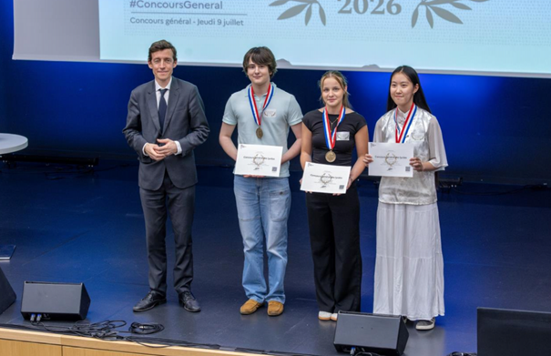 Les trois médaillés du concours général des lycées se tiennent sur une scène avec la ministre de l'éducation nationale. Ils ont une médaille au cou et montrent leur attestation de concours