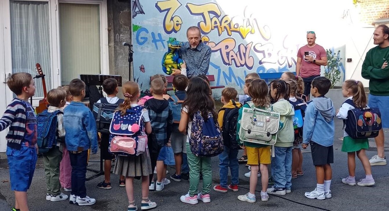 Plusieurs élèves avec leurs sacs sur le dos se trouvent dans une cour d'école et regardent une marionette en forme de dragon