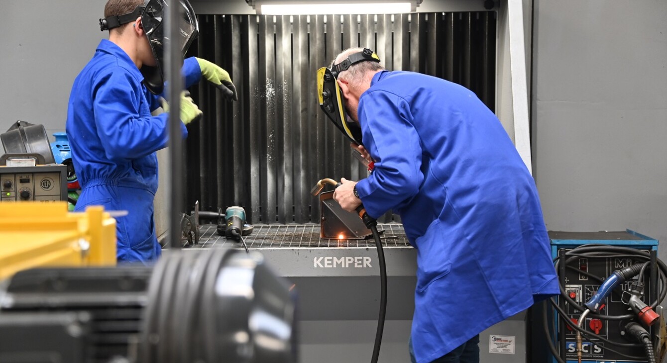 Deux personnes en bleu de travail sont penchées dans un appareil, elles soudent des pièces de métal