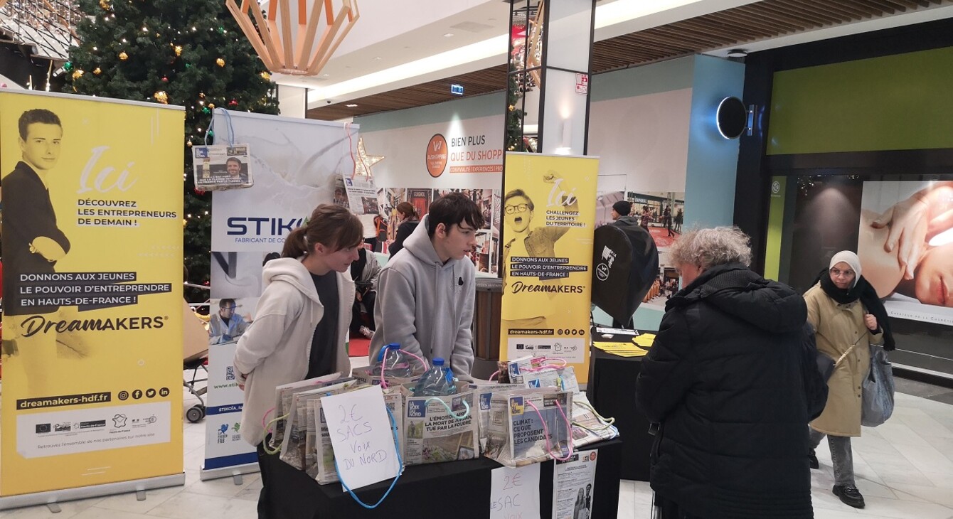 2 élèves sont dans la galerie d'un magasin et renseignent un passant sur les sacs qu'ils vendent et qui sont faits avec des journaux