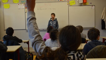 Un collègien se tient debout, au tableau, devant sa classe, une collégienne assise à sa table lève la main