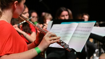 Une élève joue de la flute traversière, une partition se trouve devant elle, et d'autres élèves jouent un instrument en arrière plan 