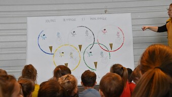 Des élèves en école primaire regarde une grande affiche sur laquelle des points colorés forment les anneaux olympiques