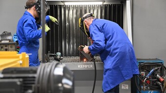 Deux personnes en bleu de travail sont penchées dans un appareil, elles soudent des pièces de métal