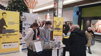 2 élèves sont dans la galerie d'un magasin et renseignent un passant sur les sacs qu'ils vendent et qui sont faits avec des journaux