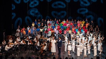 De nombreux élèves sont sur une scène, une partie des élèves chante, une deuxième partie des élèves danse et une troisième partie des élèves joue d'un instrument
