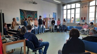 Des élèves lisent des textes dans le CDI devant un public assis autour d'eux, un enseignant les accompagne au clavecin