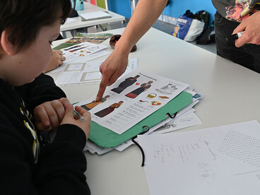 Un élève assis à sa table de classe est aidé par un enseignant sur un exercice illustrant des personnes en costumes régionaux