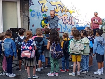 Plusieurs élèves avec leurs sacs sur le dos se trouvent dans une cour d'école et regardent une marionette en forme de dragon