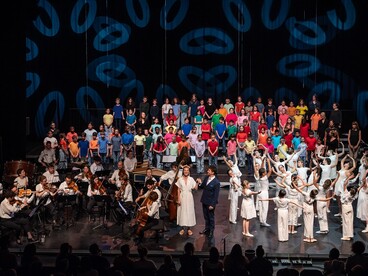 De nombreux élèves sont sur une scène, une partie des élèves chante, une deuxième partie des élèves danse et une troisième partie des élèves joue d'un instrument
