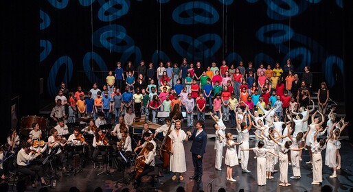 De nombreux élèves sont sur une scène, une partie des élèves chante, une deuxième partie des élèves danse et une troisième partie des élèves joue d'un instrument