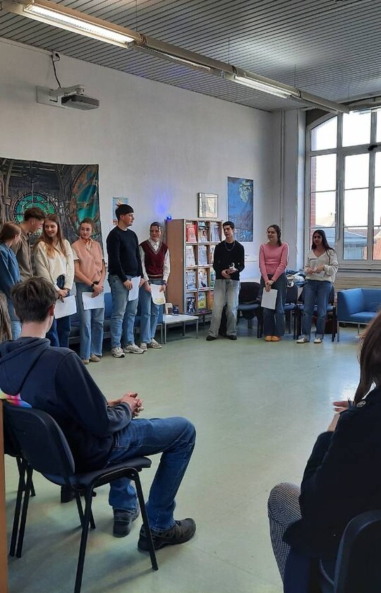 Des élèves lisent des textes dans le CDI devant un public assis autour d'eux, un enseignant les accompagne au clavecin